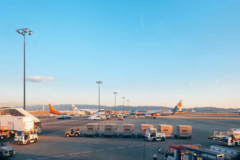 Airport ramp with ground handling vehicles and parked aircraft, lit by high mast floodlight towers across the apron