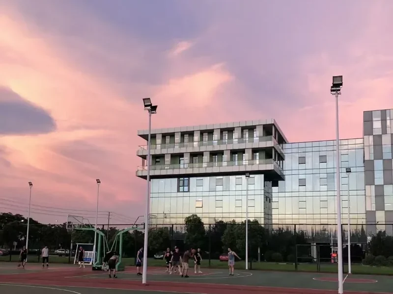 Outdoor basketball court at dusk with floodlight masts around the perimeter and apartment blocks in the background
