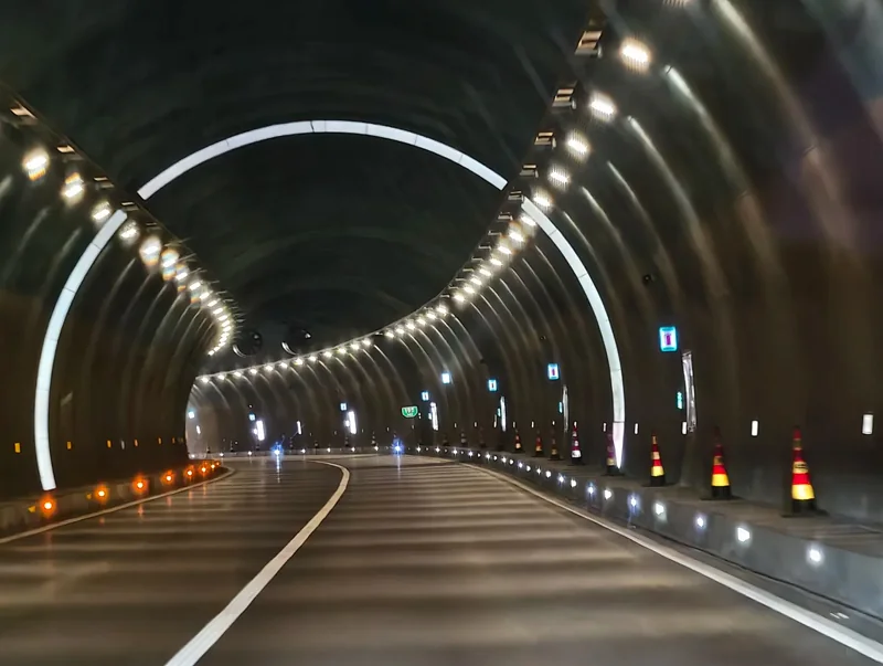 Arched road tunnel lit by continuous rows of LED tunnel lighting along the crown, with reflective lane markers and traffic cones on the carriageway